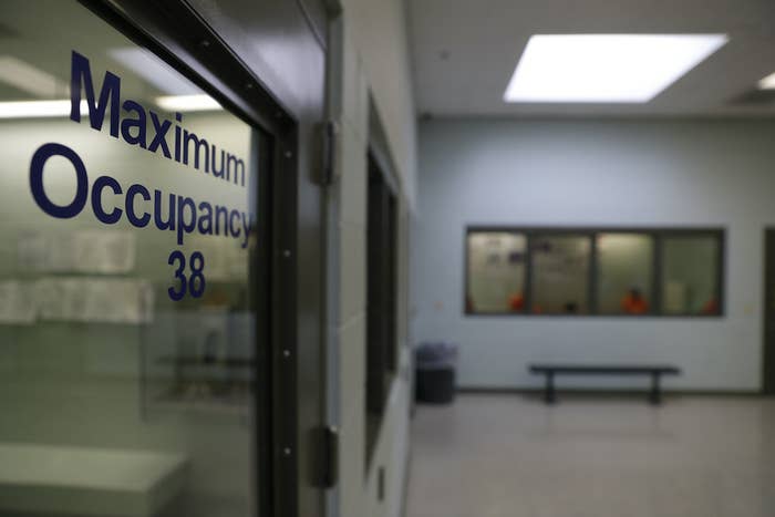 People wearing brightly colored jumpsuits are visible through a window in an ICE processing center