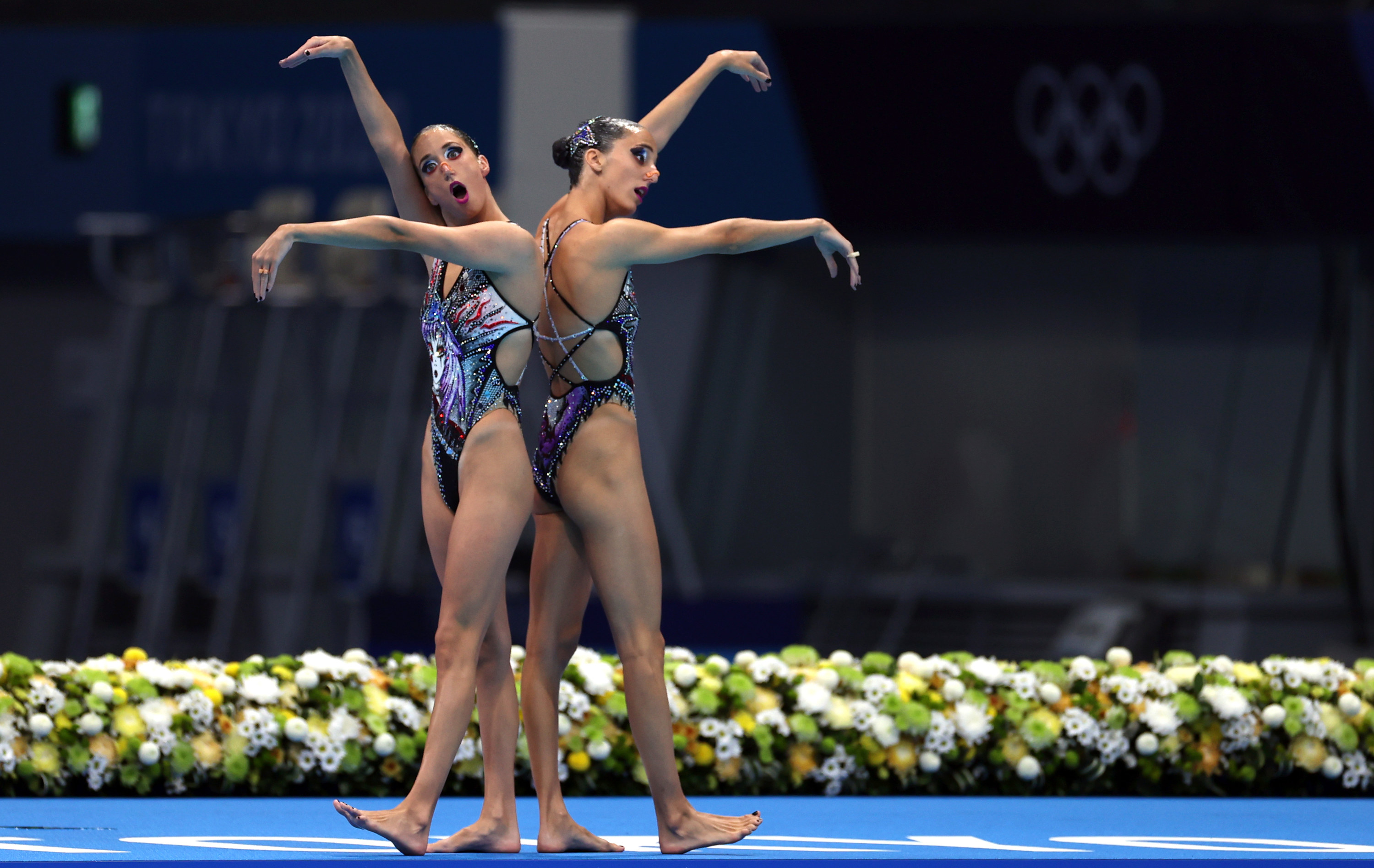 Two women swimmers stand back to back on a mat with their arms stretched before them in a performance