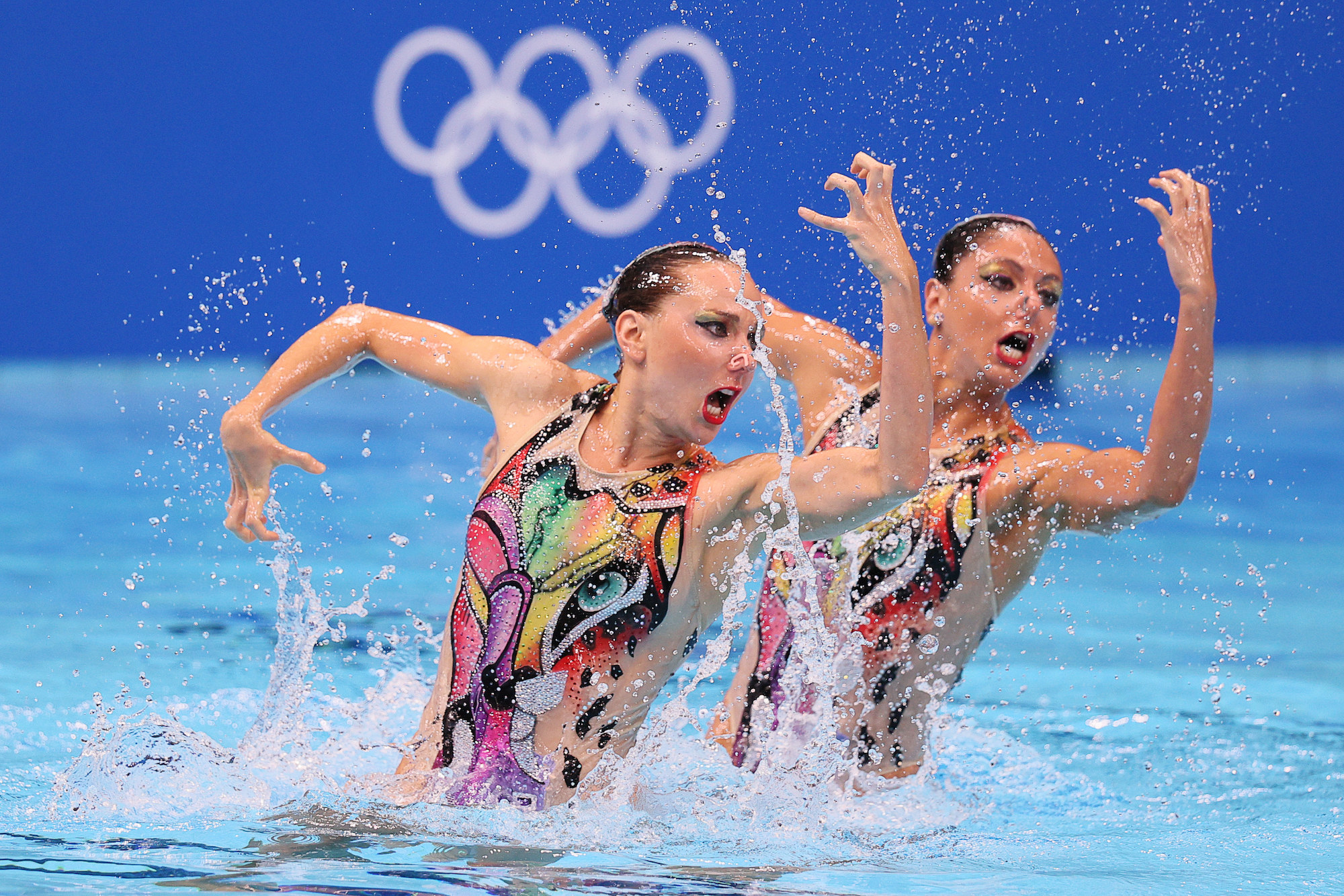 Two women swimmers perform in the pool at the Tokyo Olympics with their arms in front of and behind them