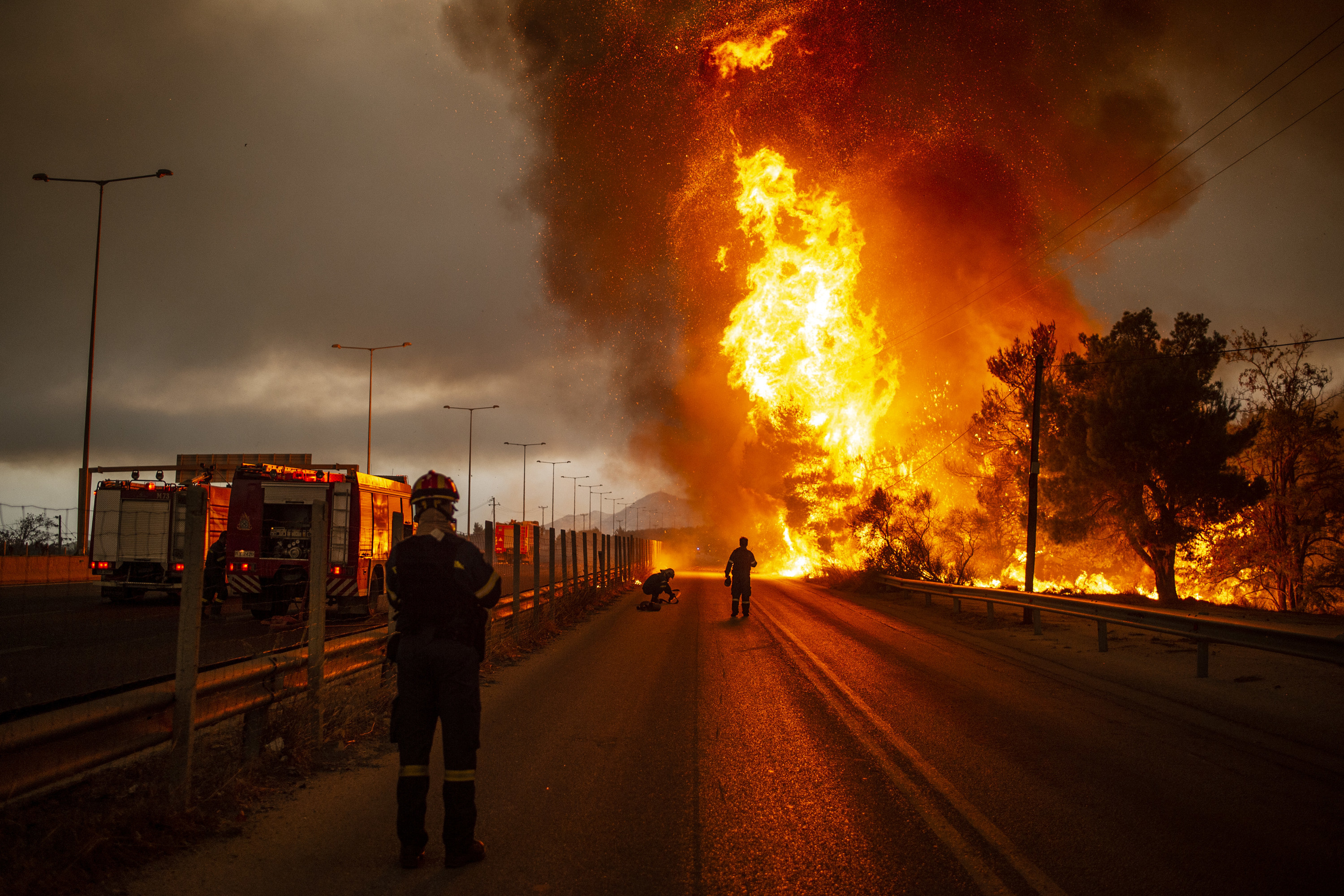 A firefighter stands at the side of a main road as flames shoot up into the sky several meters in front of them