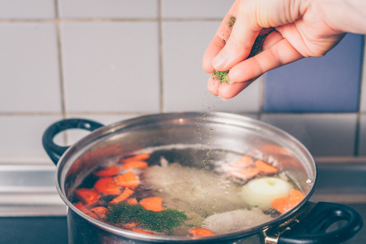 Someone adding seasoning to a pot of chicken soup.