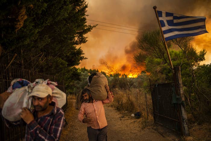 Two men carrying their belongings on their shoulders as they walk by a fence with the Greek flag as fire rages in the background