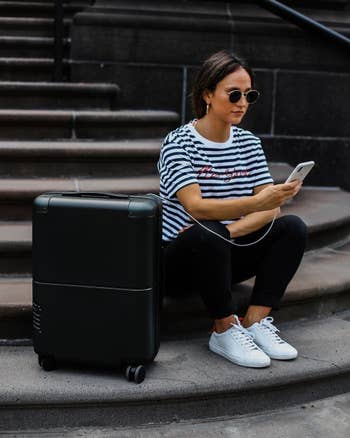 model sitting next to the black suitcase while charging a phone from it