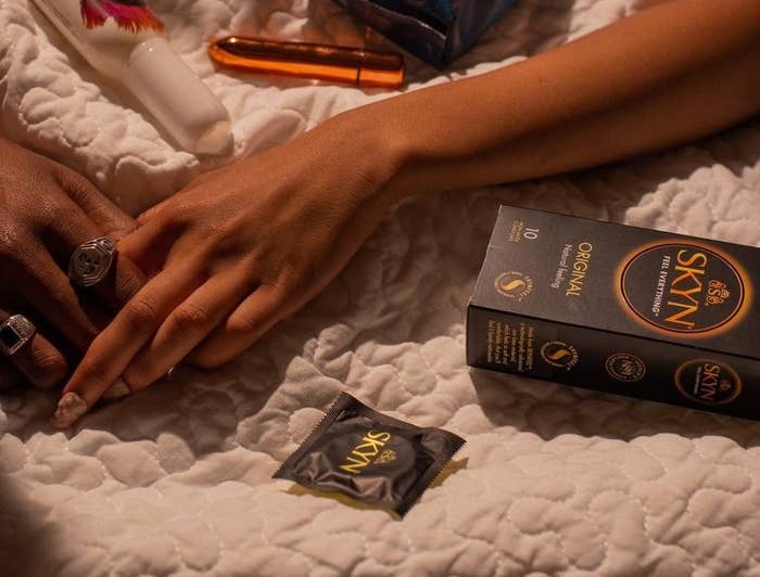 Models holding hands on bed surrounded by various sexual wellness products