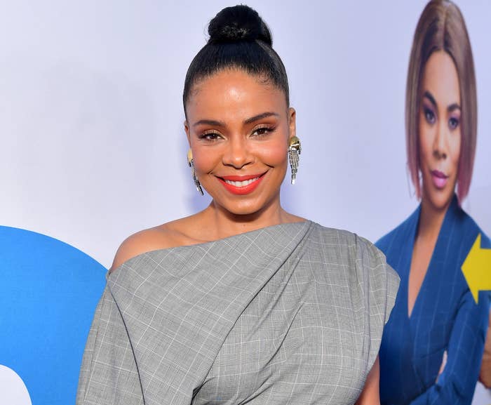 Sanaa smiles while wearing her hair in a bun and an off the shoulder grey and white plaid dress