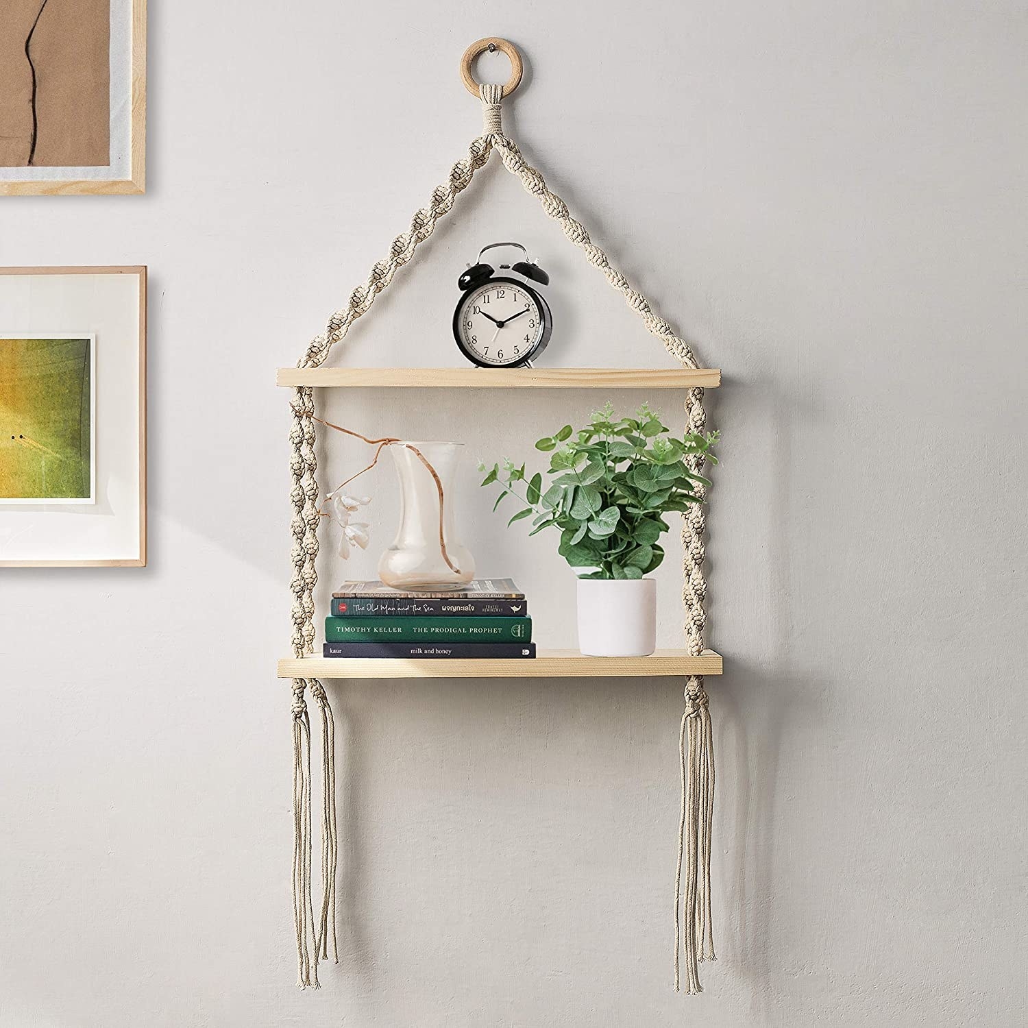 The white macrame wall hanging shelf with books, a potted plant and a clock placed on it