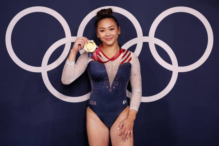 Sunisa holding up her gold medal as she poses in front of the Olympic rings logo