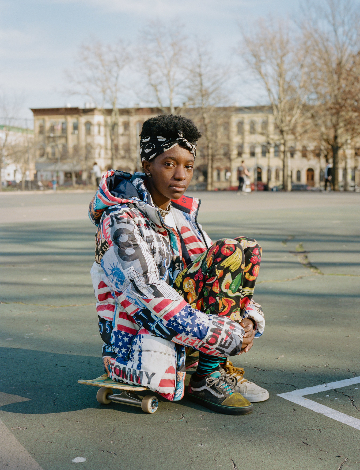 A woman sits on a skateboard on a playground with a row of brownstones in the background