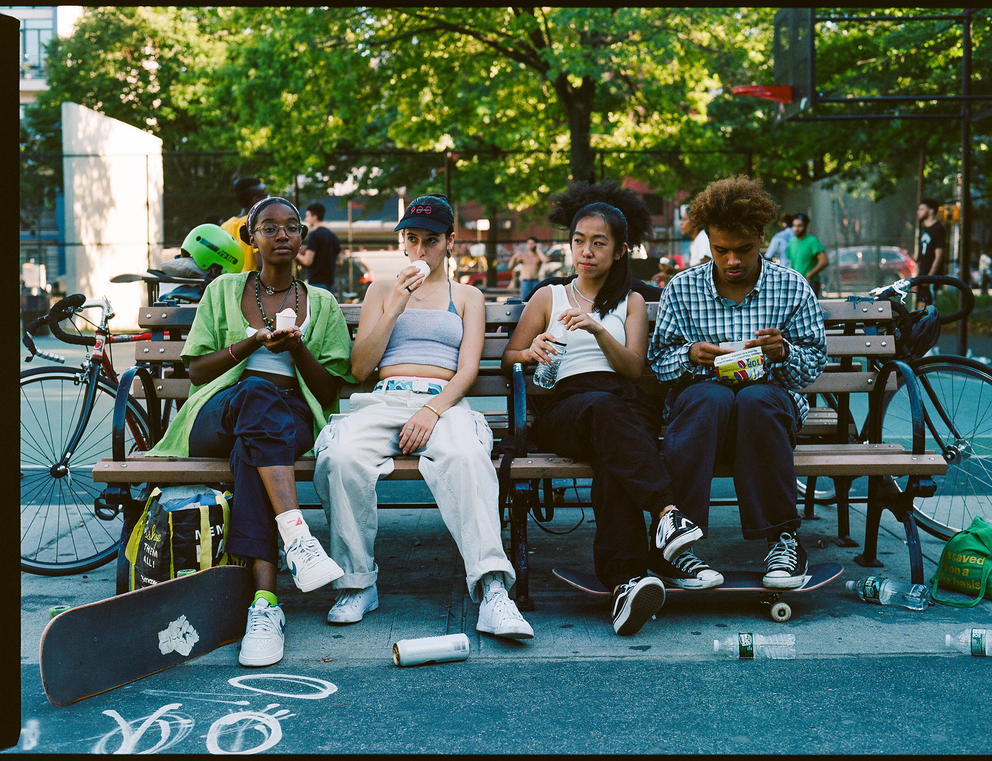 A group of women sitting on a park bench eat a snack with skateboards at their feet 