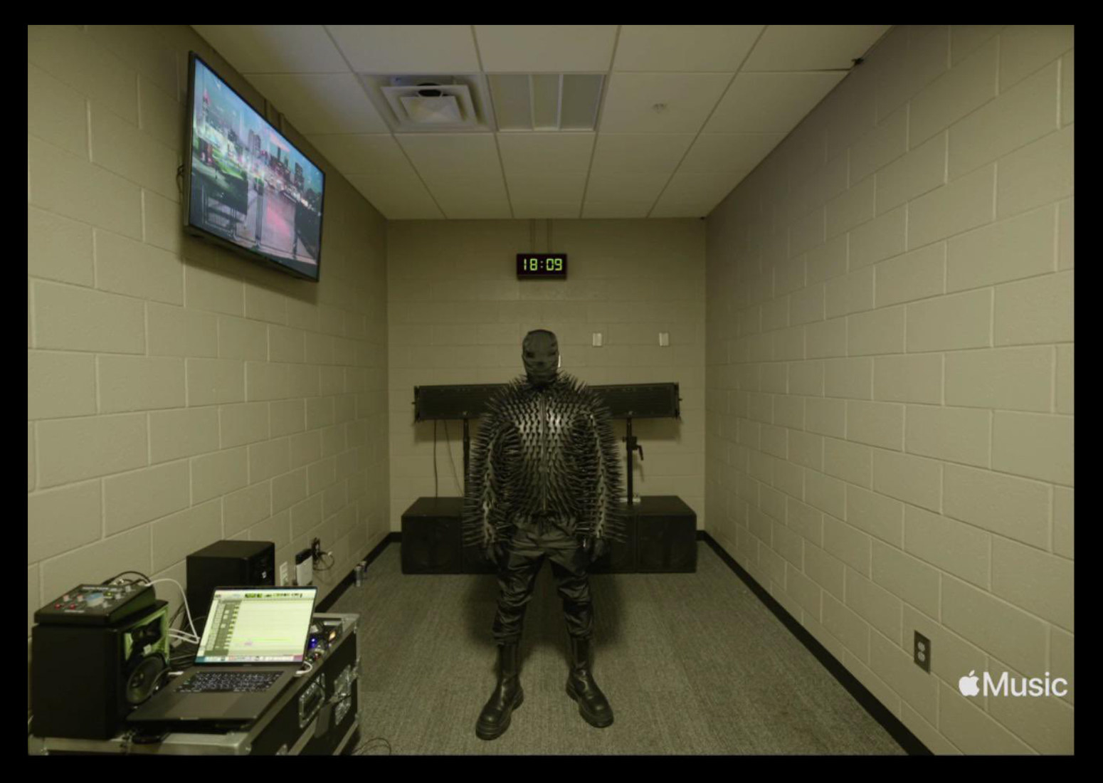 Kanye wears a spike-covered shirt, pants, boots, and a full face covering, standing in a concrete-lined room