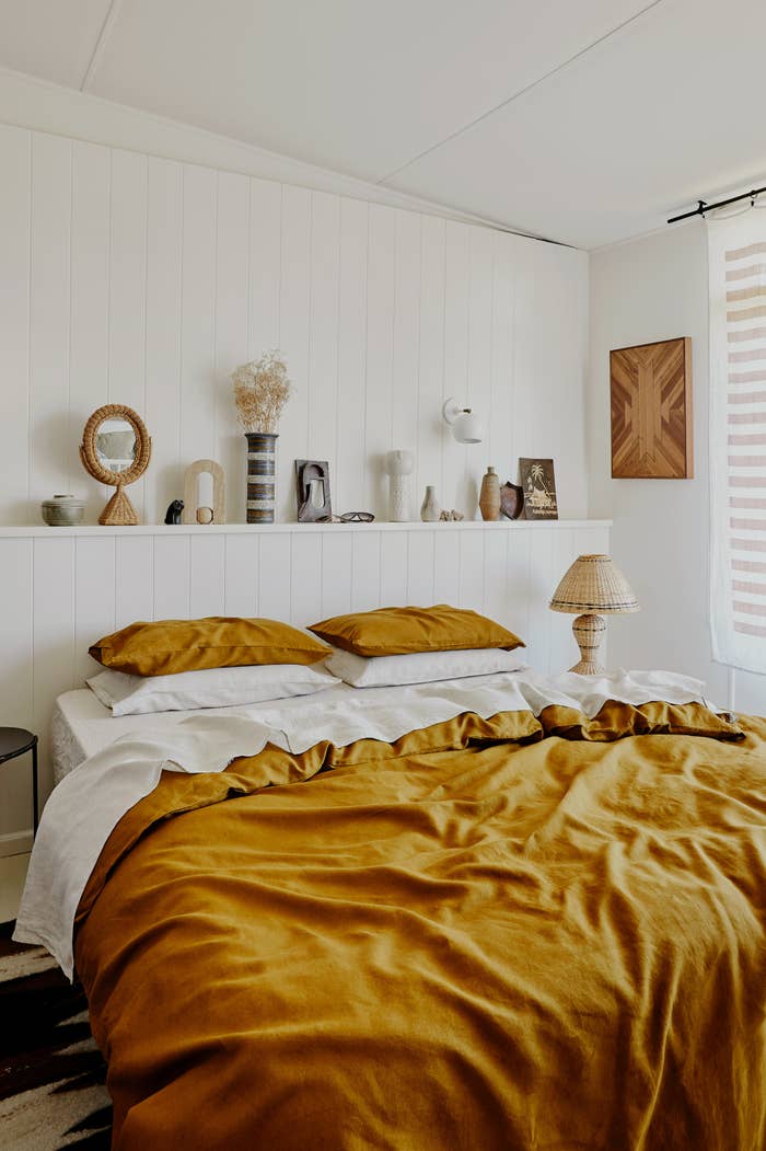 Bed without a headboard dressed with mustard and white linen bedding including a fitted white linen sheet, four pillows of which two are mustard and two are white, a mustard duvet and a white flat sheet underneath.