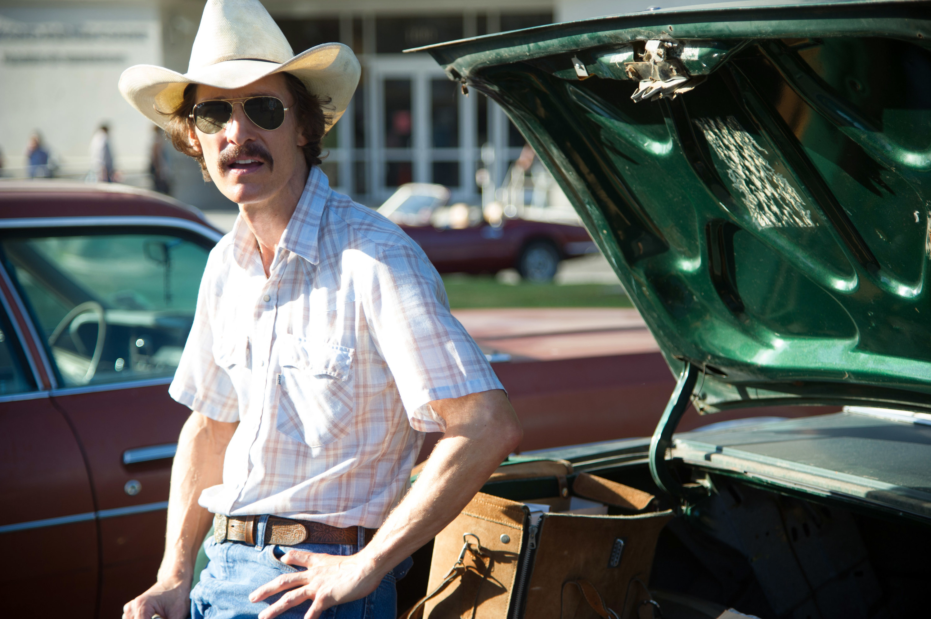 Matthew as Ron wearing a cowboy hat and leaning against a car with his hand on his hip