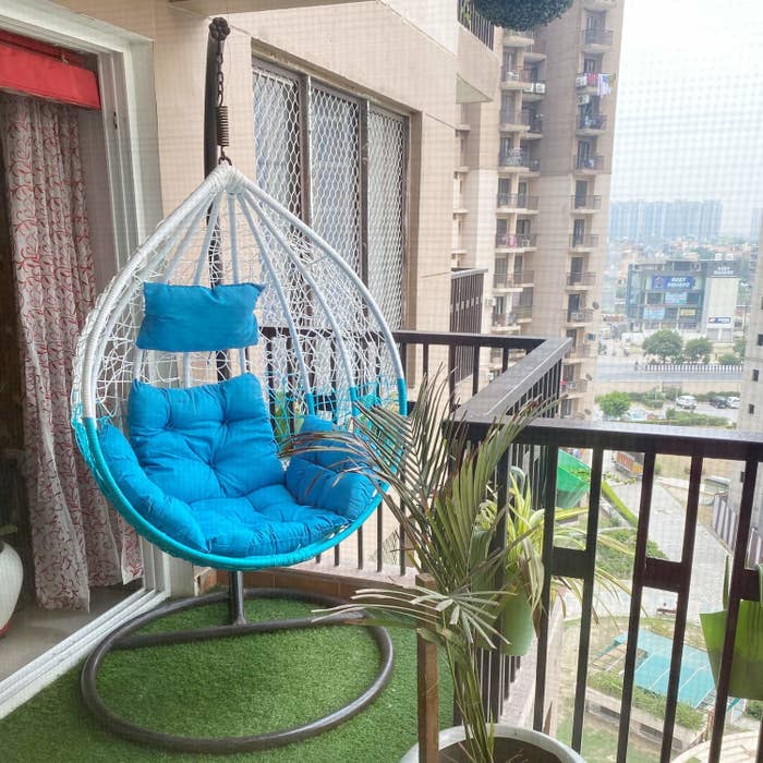A blue and white pod-shaped swing chair with a stand kept on a balcony with blue cushions