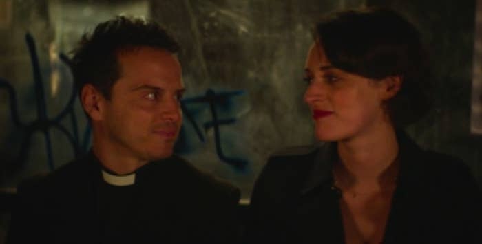 Hot Priest and Fleabag look at each other with soft smiles while sitting at a bus stop covered in graffiti