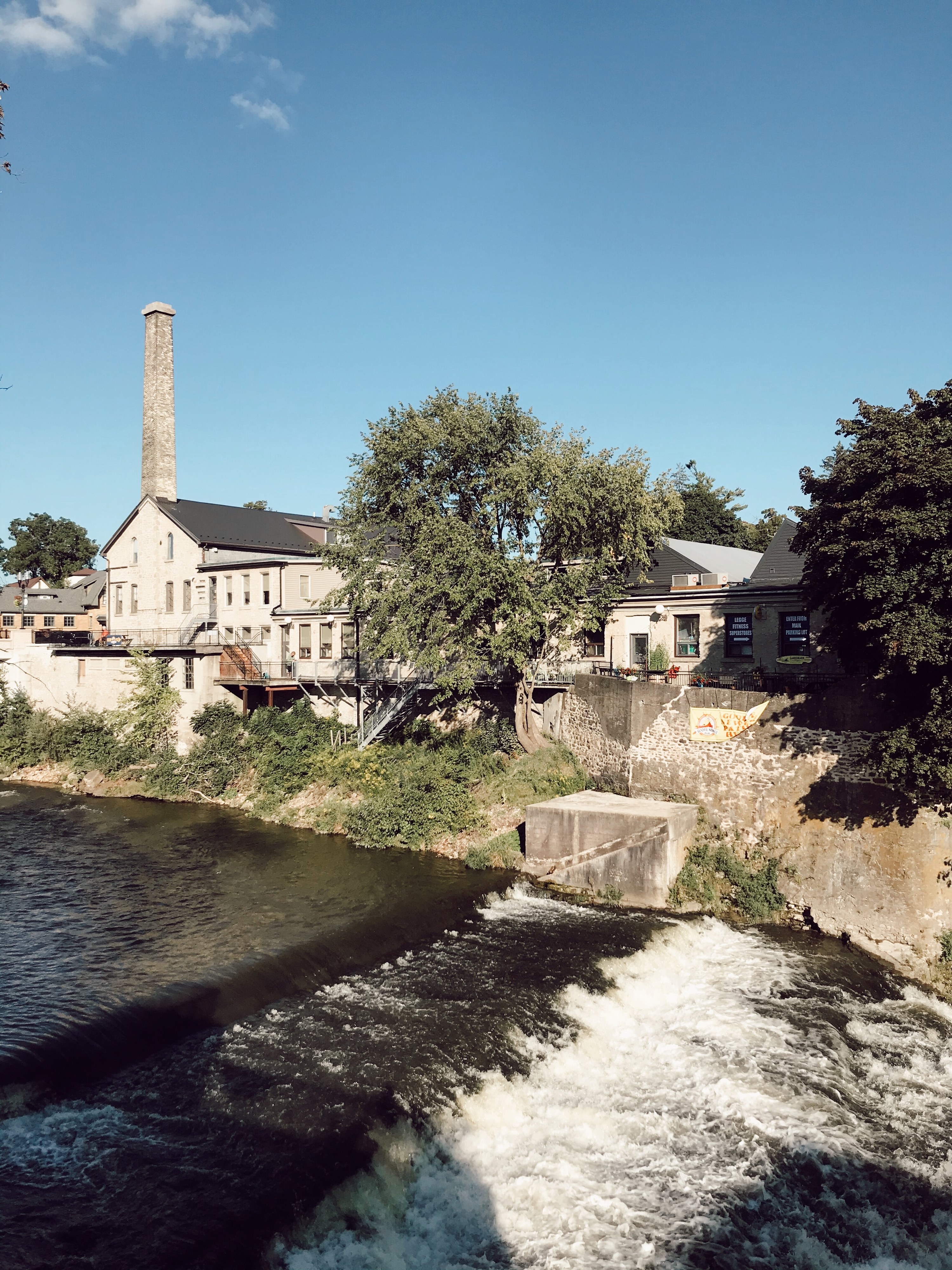Photograph of a fast river, rushing along a tall stone wall, where several European-style stone buildings sit. One of the stone buildings has a large chimney