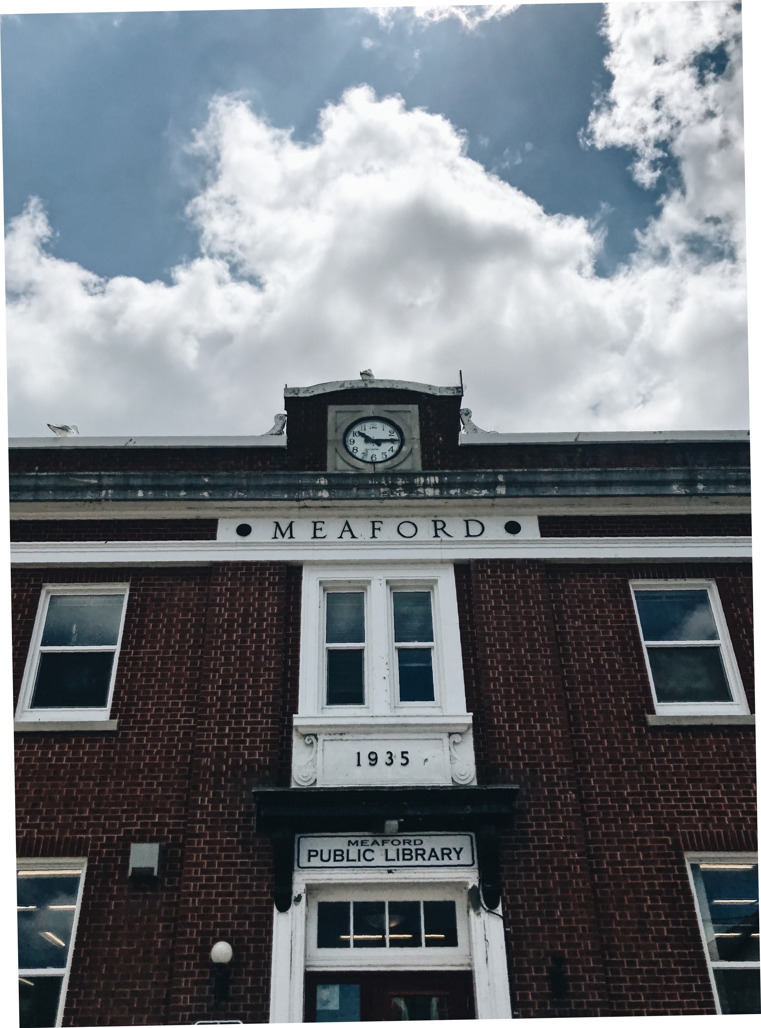 A photograph of the front of the old Meaford Library. It&#x27;s a red brick building with a sign above the door that reads &quot;MEAFORD PUBLIC LIBRARY&quot; and a clock at the very top of the front wall