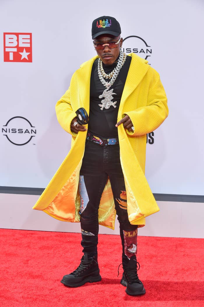 DaBaby on a red carpet in dark jeans, a long brightly colored jacket and combat boots