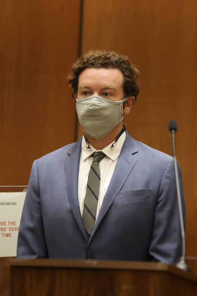 Danny Masterson in a suit and tie, standing in a courtroom and wearing a mask
