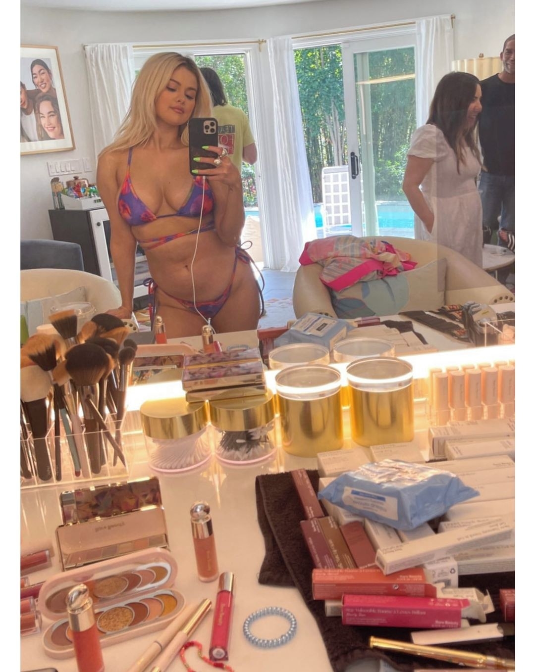 Selena taking a selfie at a mirror, wearing a bathing suit, with a table with makeup and applicators in the foreground