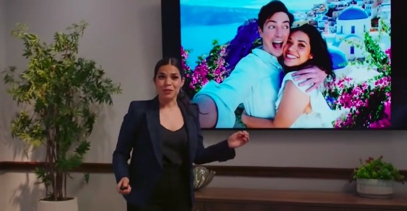 Amy stands in a business suit in front of a screen showing a photo of her and Jonah embracing in Greece