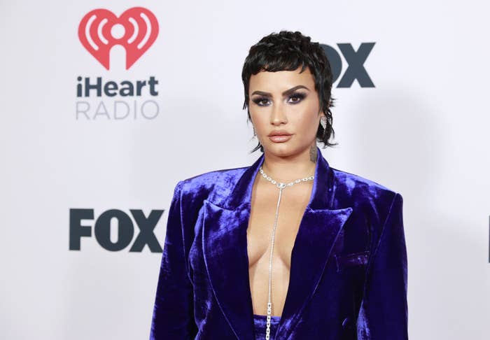 Demi Lovato posing on a red carpet in a open velvet jacket with matching pants, no shirt, and a diamond necklace