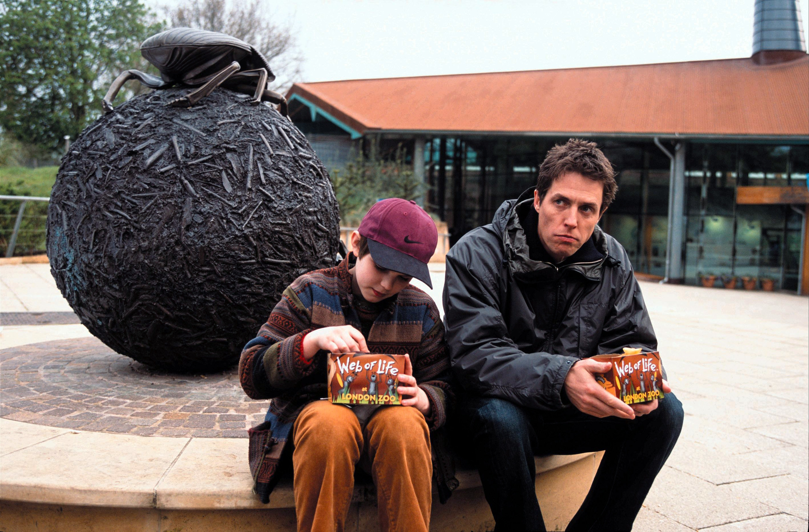 Nicholas Hoult and Hugh Grant sit outside in front of a sculpture