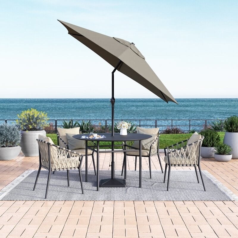 the umbrella tilted over a round black table and woven chairs on a patio with the ocean in the background