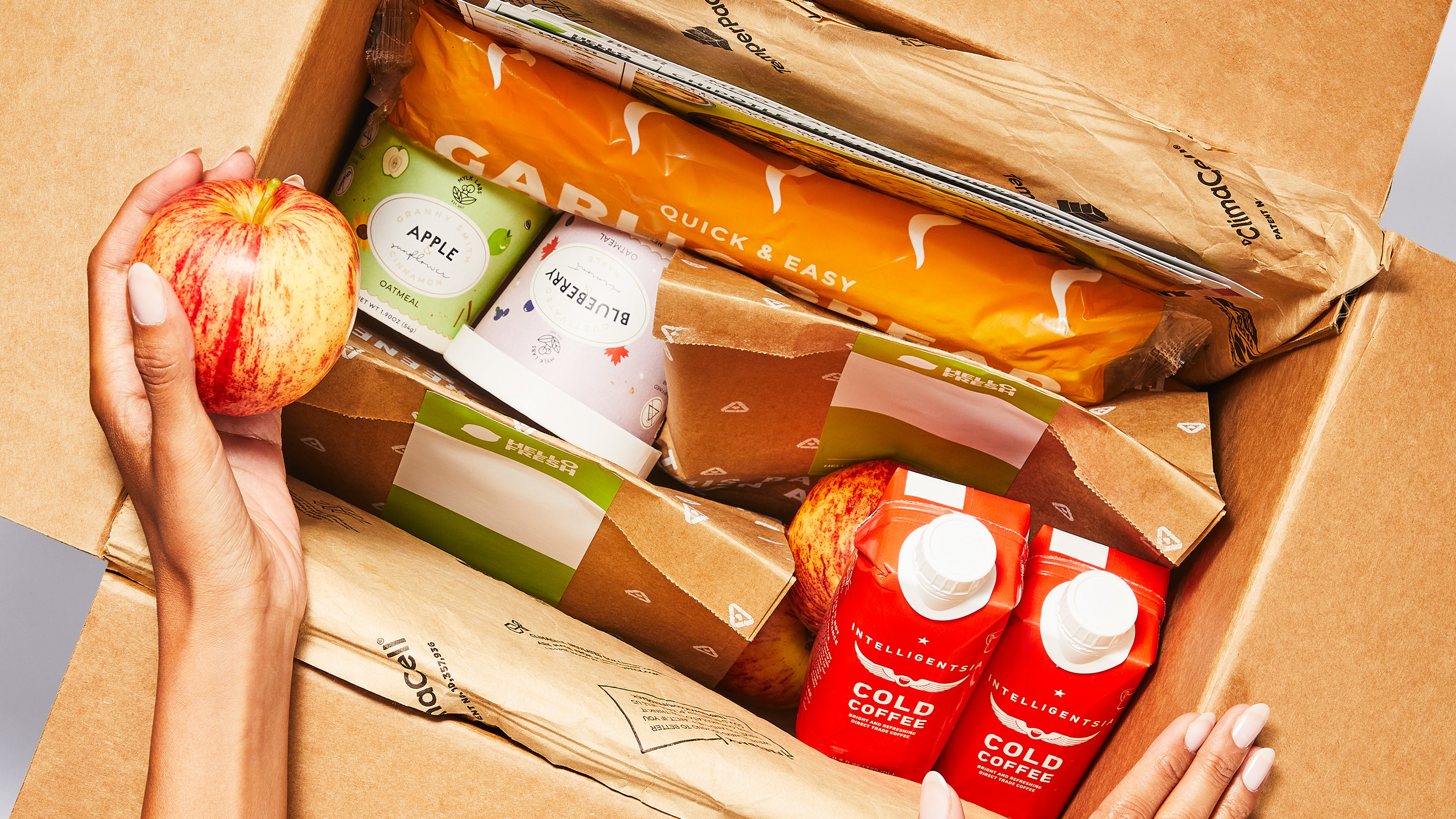 Hands are shown opening a cardboard box filled with produce and boxed coffee