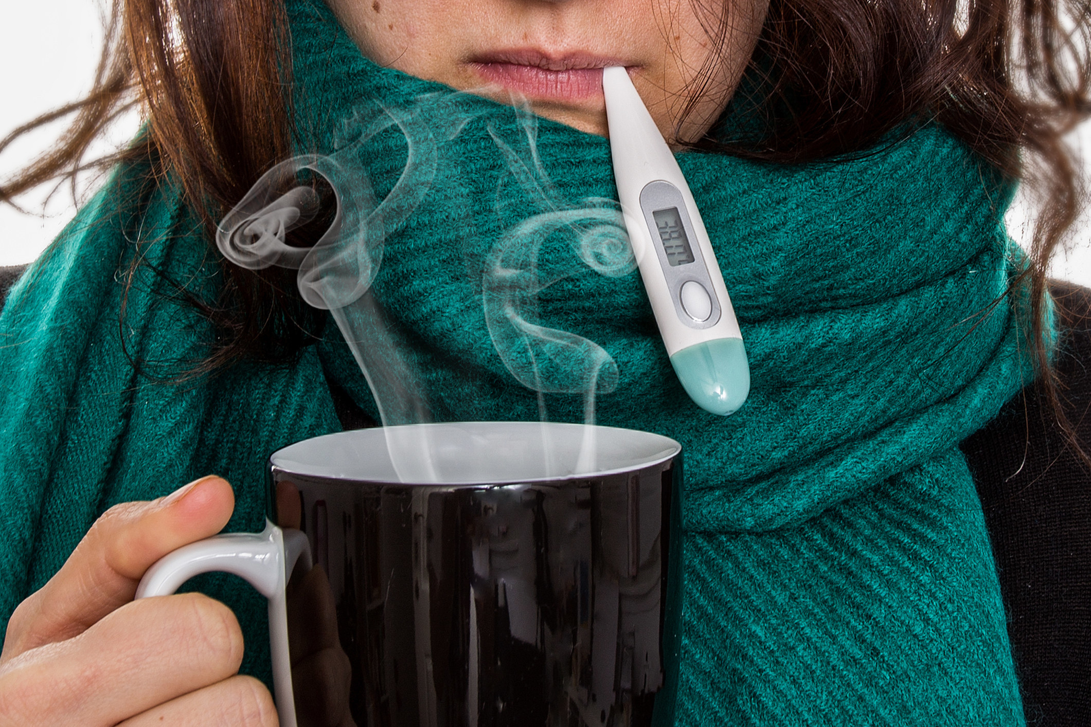 A woman with a cold holding a cup of hot tea and a thermometer in her mouth