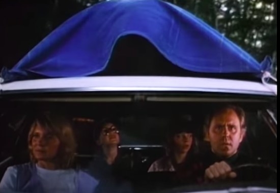 The Henderson family sit in their car with something strapped under a blue tarp to the roof of the car