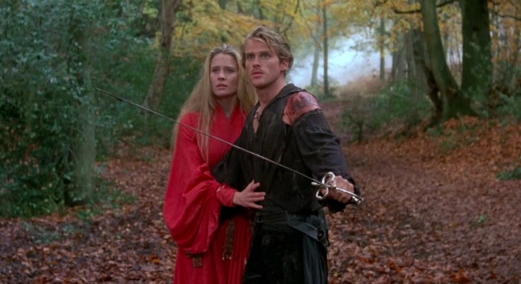 Wesley stands in front of Buttercup with his arm in front of her protectively; his other hand holds a sword; they stand in a forest