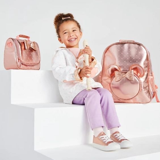 A child model sitting next to the rose gold lunch tote and matching backpack