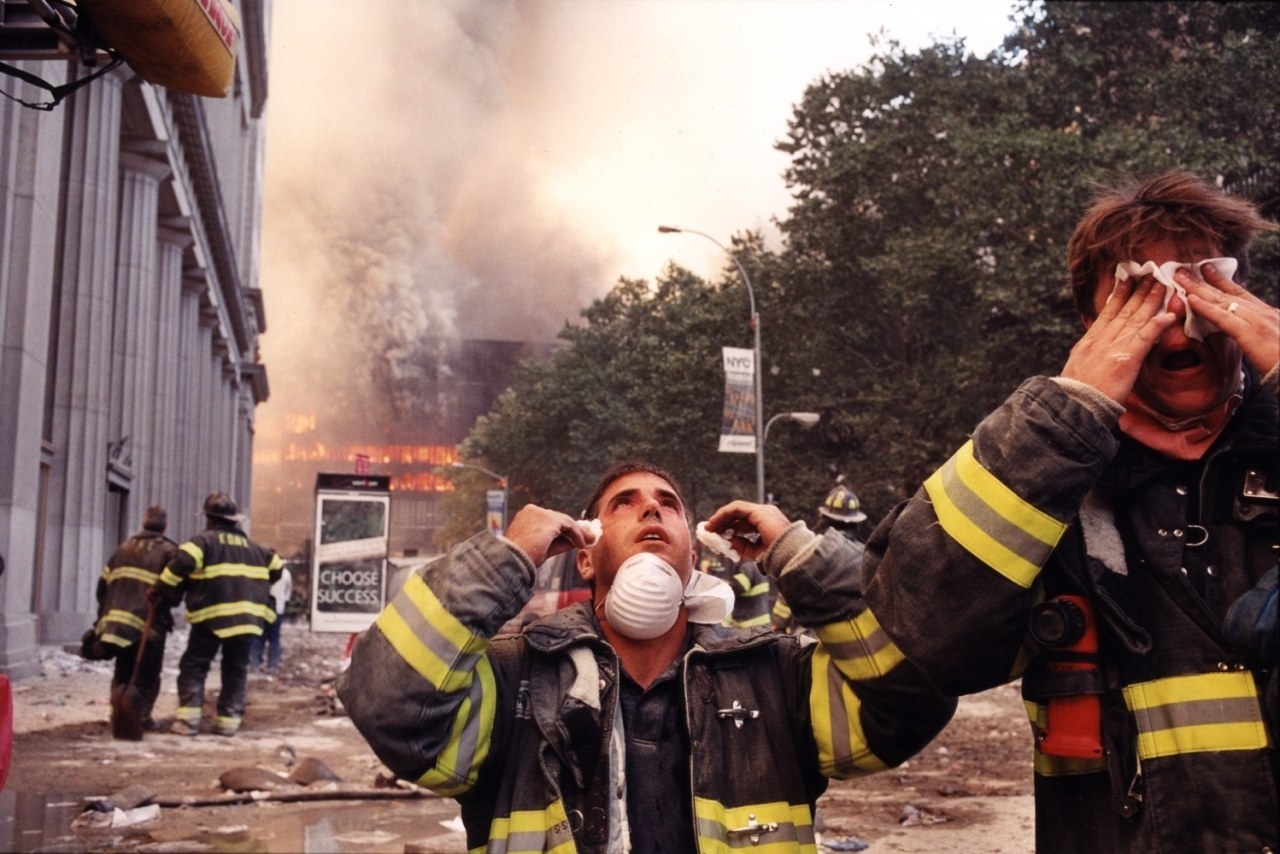 Firefighters wipe their eyes as buildings burn in the background