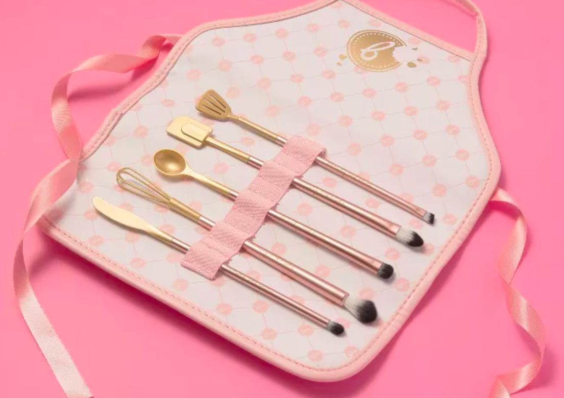five brushes inside the lots of a makeup brush roll that&#x27;s shaped like a pink and white polka-dotted apron. the brushes have handles shaped like different cooking utensils including an egg beater, butter knife, spatula, and the scraper spatula