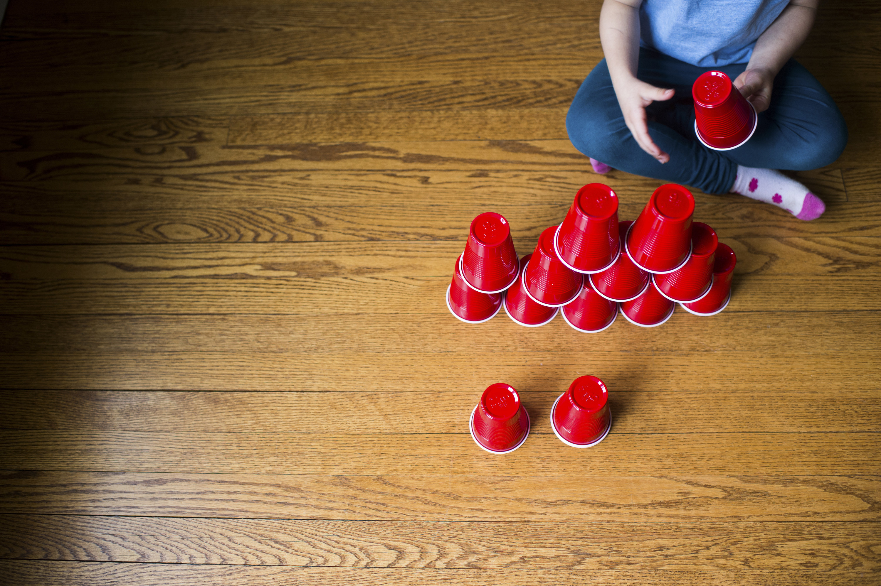 Child stacks plastic cups into a pyramid shape on a wooden floor.