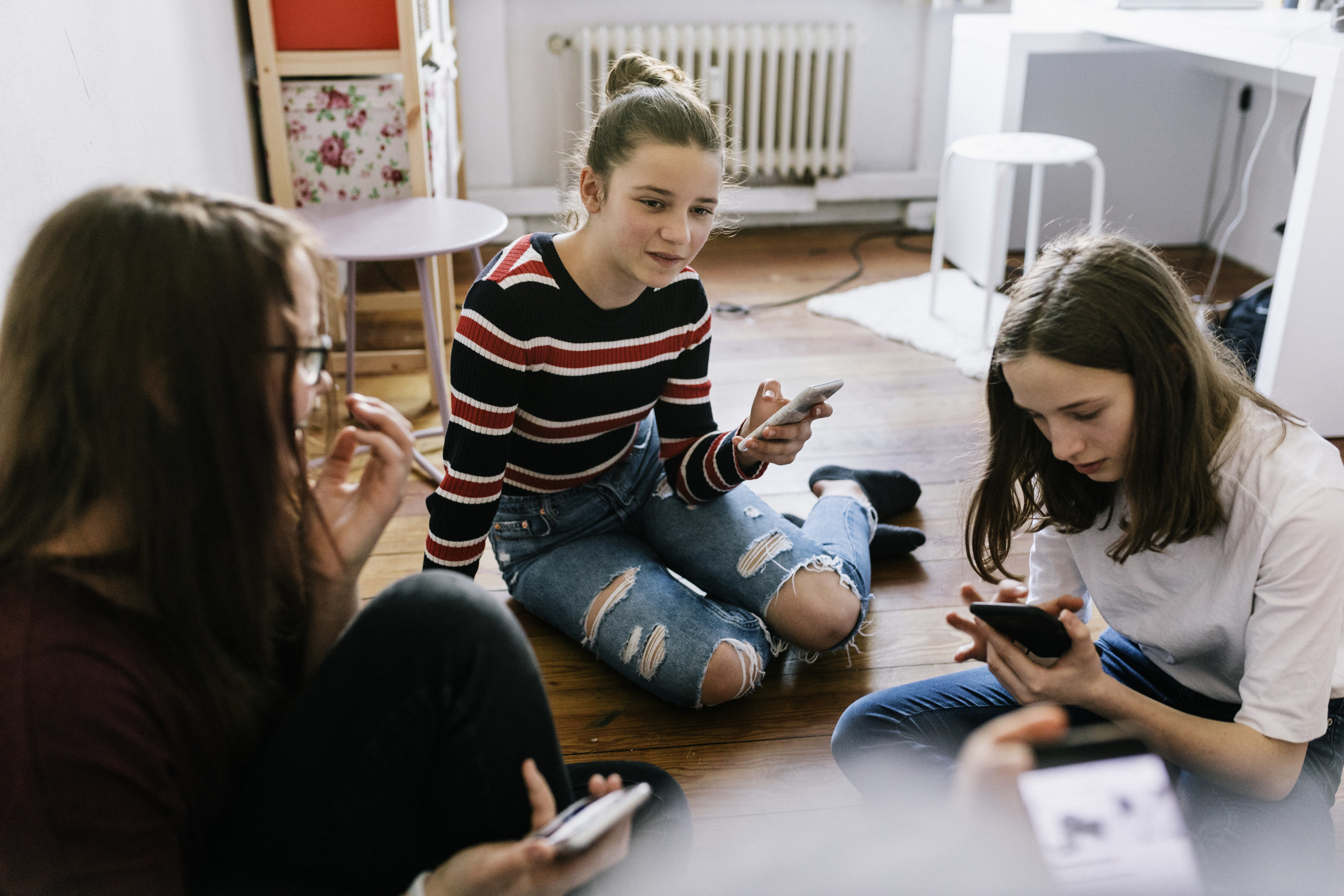 Pre-teen girls look at their phones and talk to each other in the middle of a bedroom.