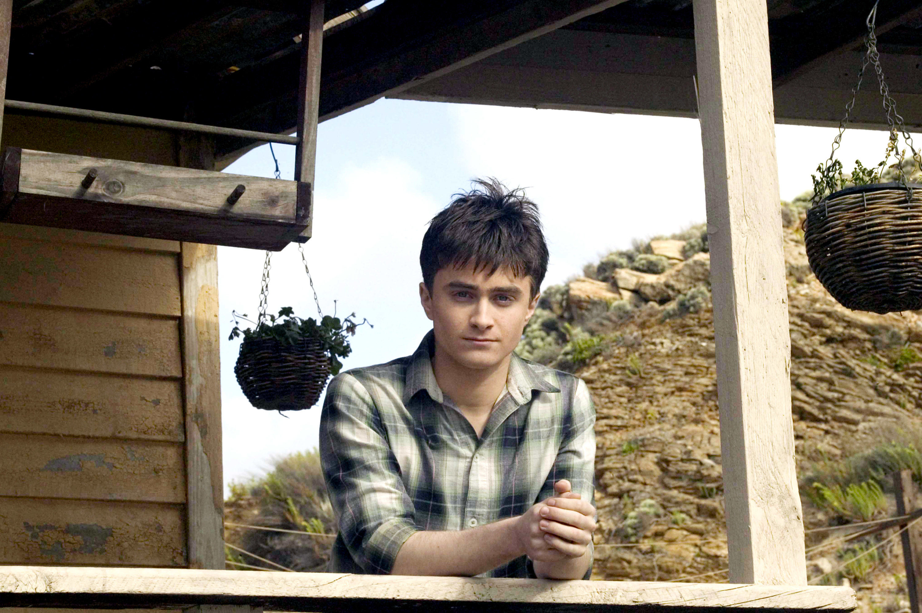 Daniel Radcliffe leans against the railing of a beach house wearing a light colored flannel shirt
