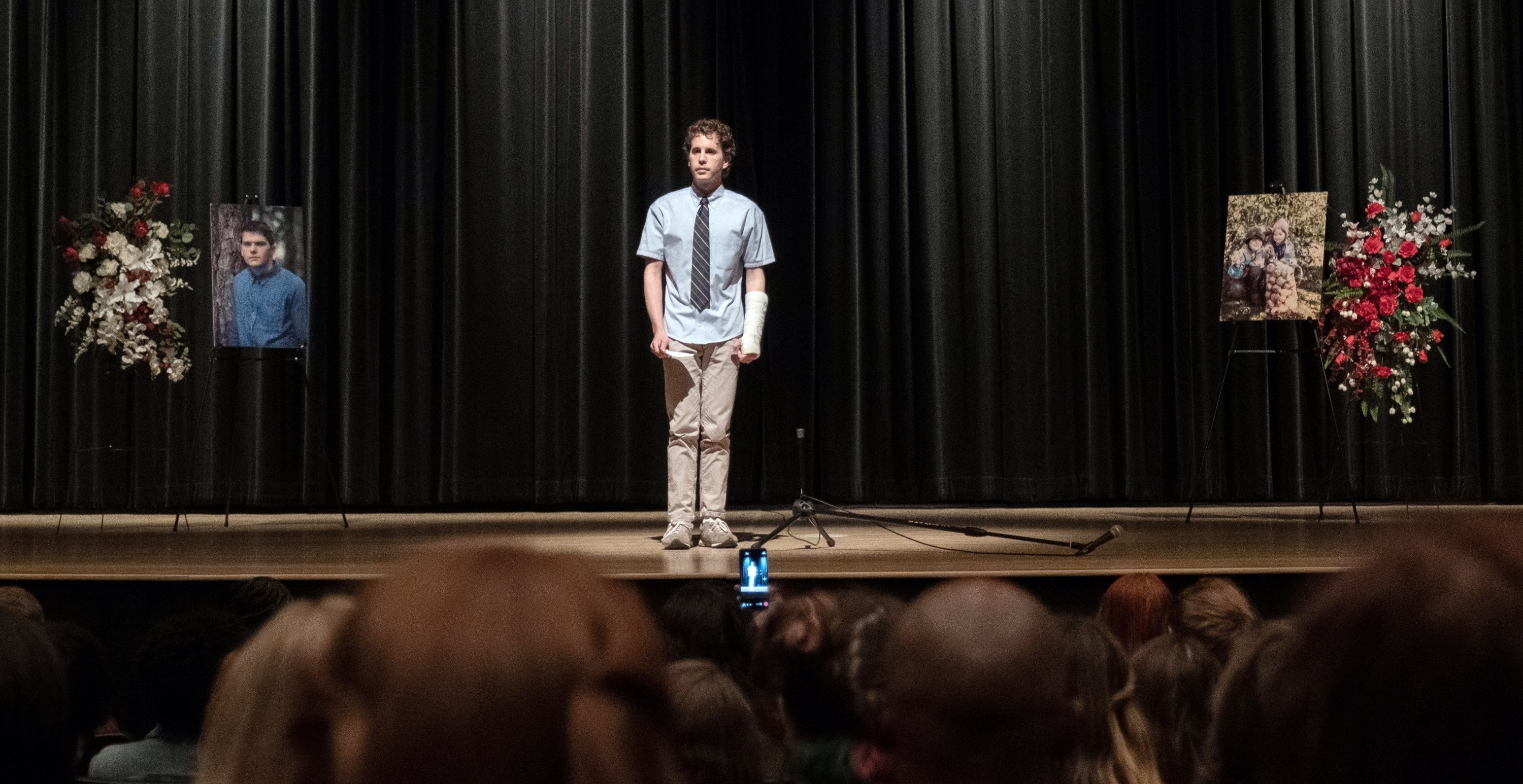 Evan Hansen standing at the mic in a school auditorium with pictures of a fellow student who has died off to the sides