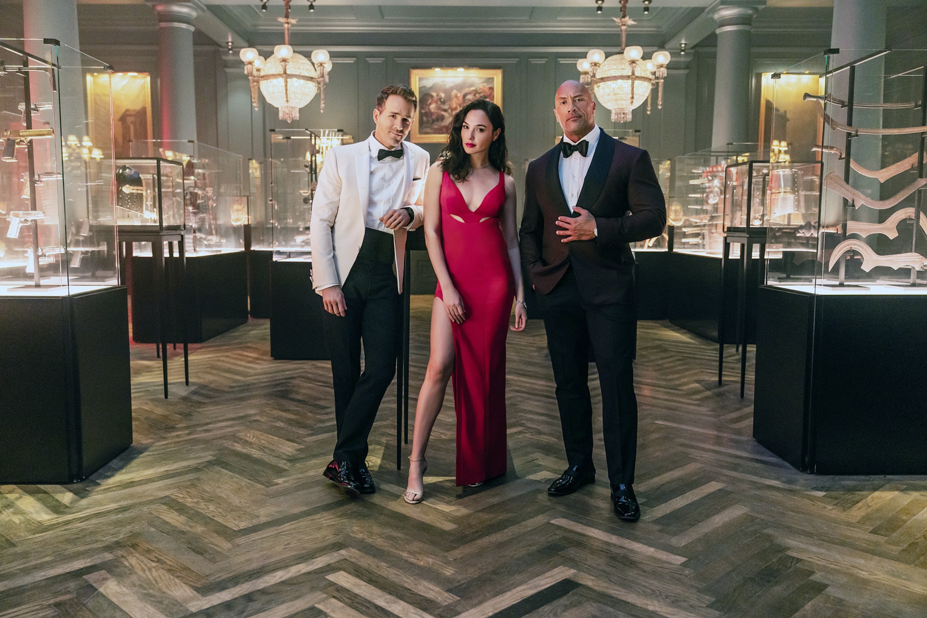 Dwayne Johnson, Gal Gadot, and Ryan Reynolds wearing evening garb as they stand in a room full of weapons in glass cases