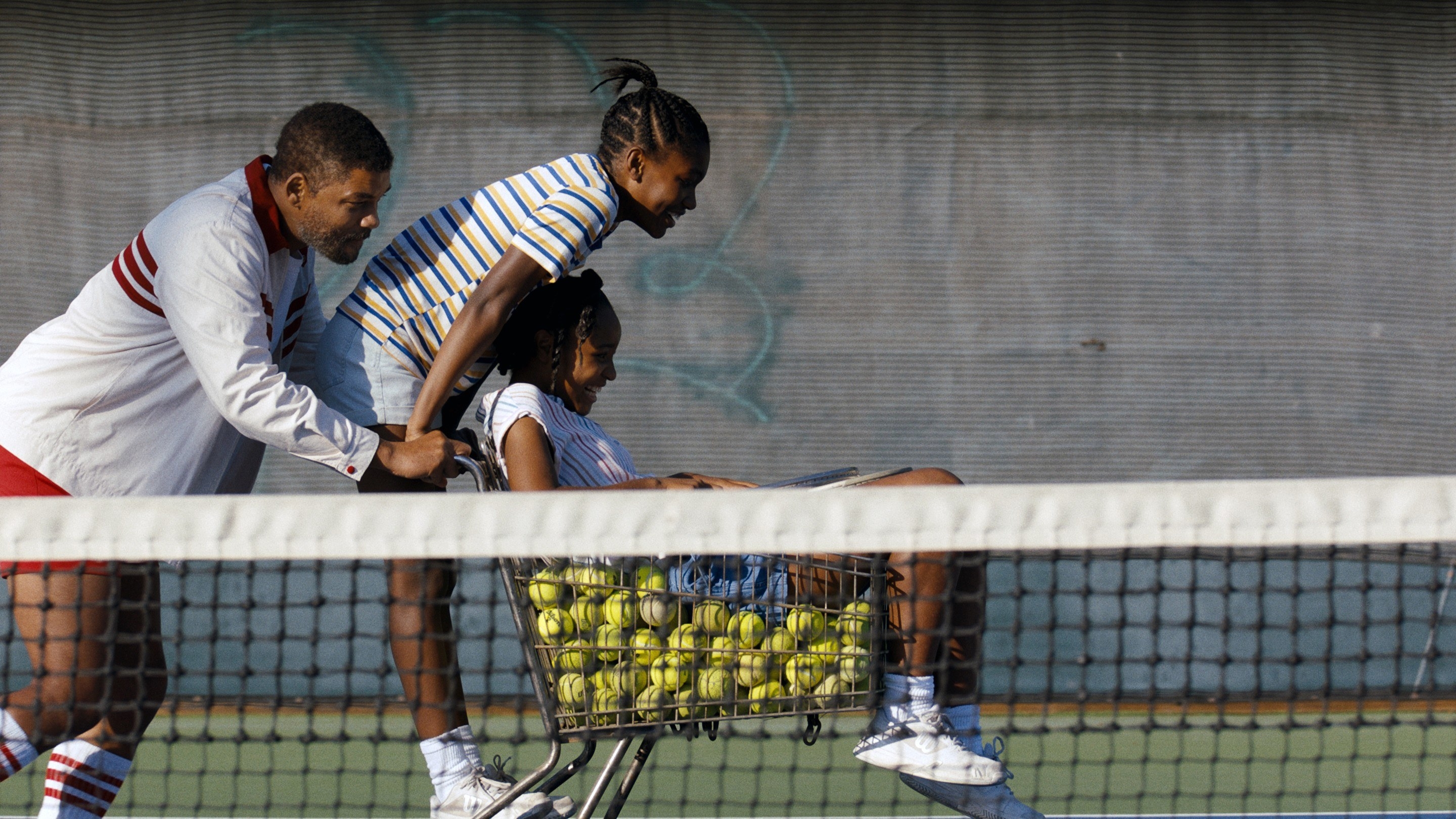 Richard pushing Venus and Serena across a tennis court as they ride on a shopping cart filled with tennis balls
