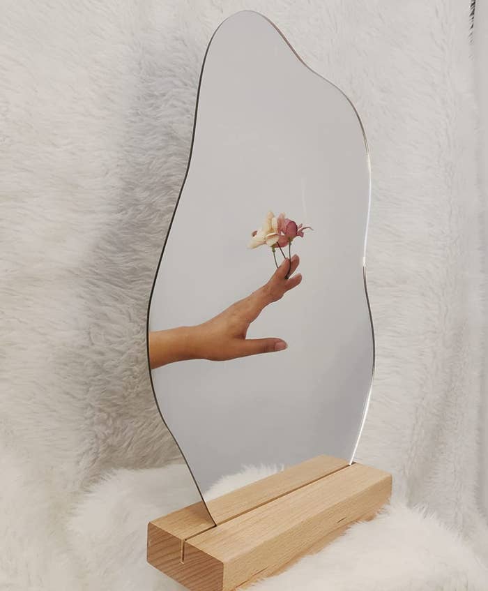 An irregular vanity mirror on a wooden stand, with a flower in someone&#x27;s hand in the reflection.
