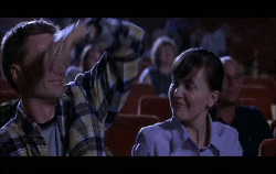 A man puts his arm around his date&#x27;s shoulders, an older woman cracks her knuckles, and a person in a &quot;Scream&quot; costume puts their feet up on the movie theater seats in front of them