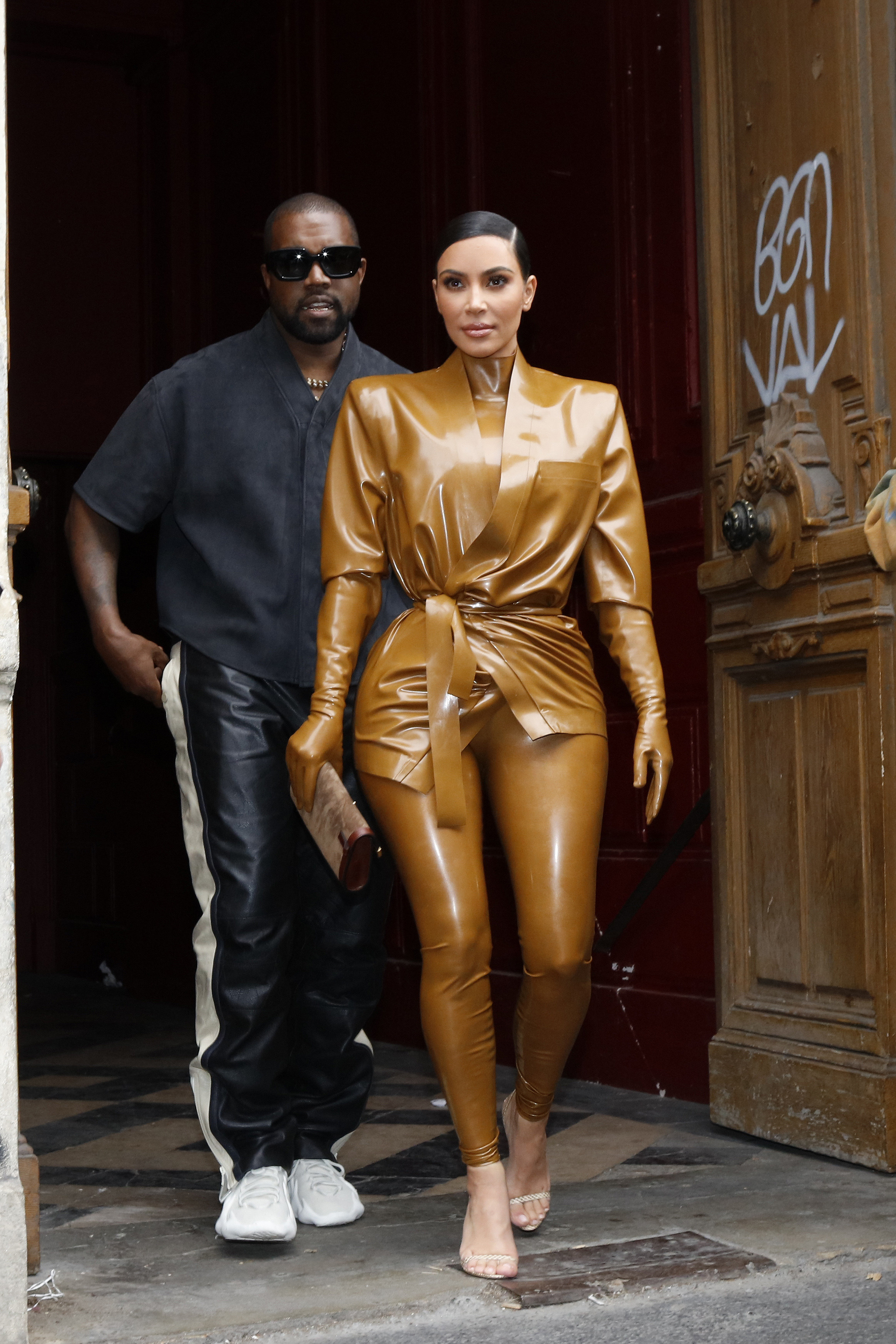 Kanye on the left and Kim, wearing a latex bodysuit with gloves, walking out of a building together