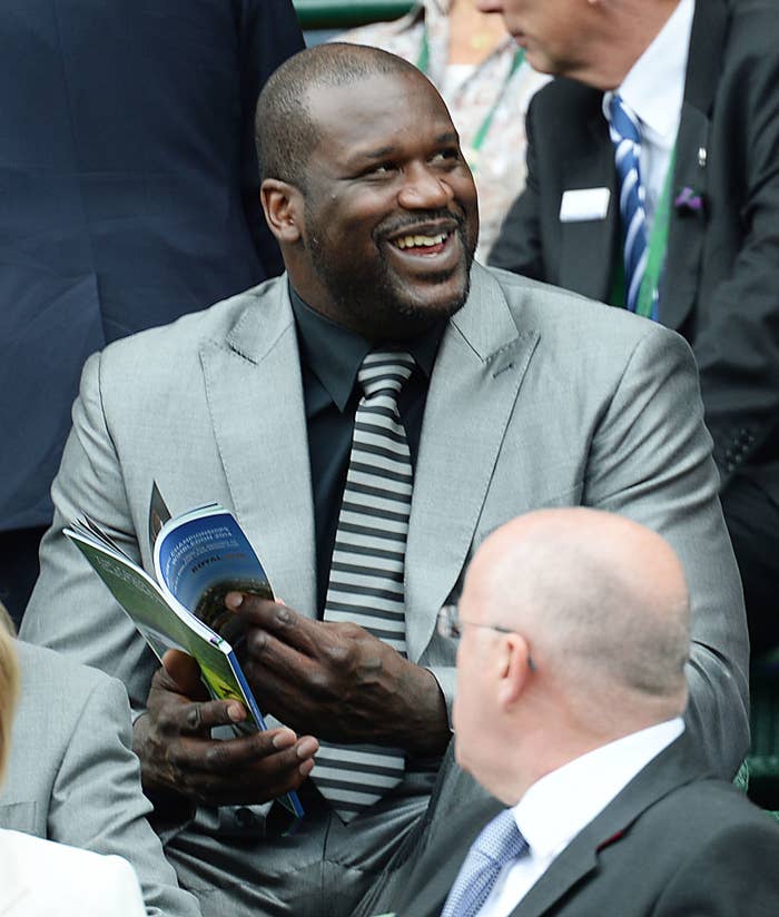 Shaq wearing a suit smiling while looking up and away from a book in his hand