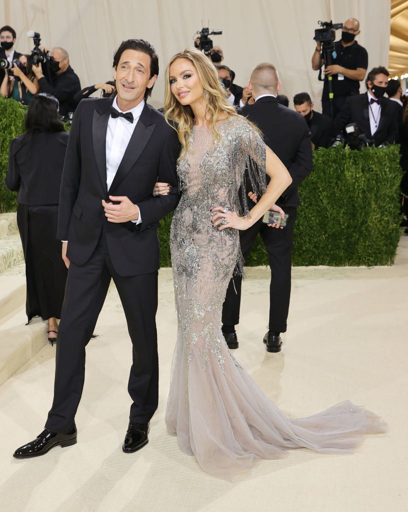 Adrien Brody wears a dark suit and Georgina Chapman wears a floor length gown with sparkles on the bodice and a long train