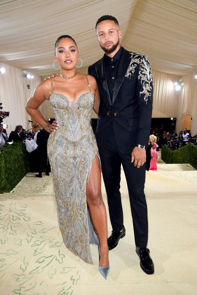 Stephen Curry wears a dark suit with a glittery pattern on the shoulder and Ayesha Curry wears a floor length thin strap glittery gown with a slit up her thigh