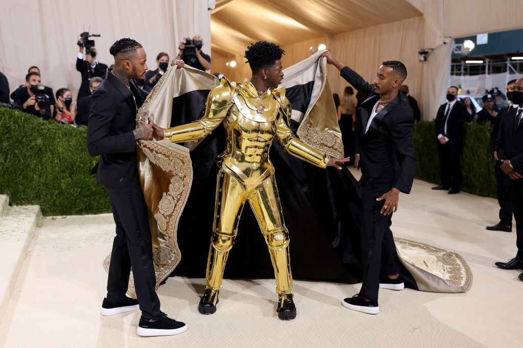 Lil Nas X&#x27;s cape is taken off by two men to reveal his gold plated suit of armor