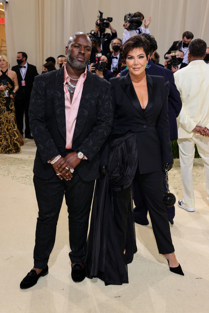 Kris Jenner  wears a dark suit with a short train wrapped around her waist and Corey Gamble wears a brightly colored button up shirt under a dark blazer and matching slacks