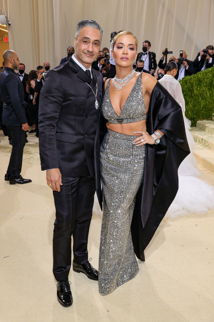 Taika Waititi wears a dark suit and Rita Ora wears a floor length sparkly skirt and matching cropped tank top