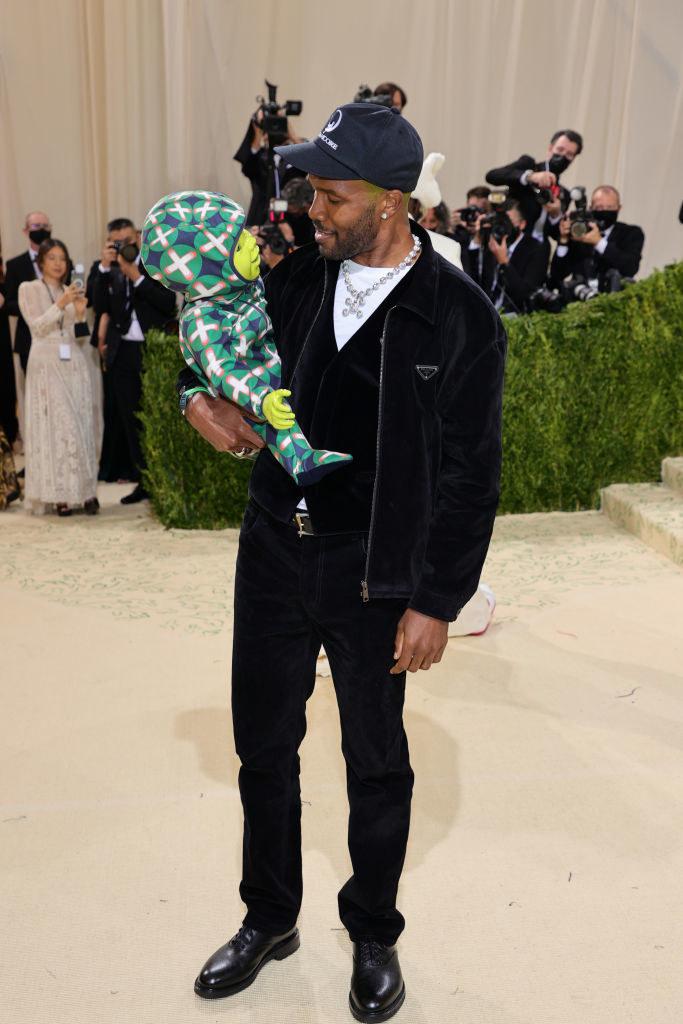 Frank Ocean wears dark slacks, a matching v-neck shirt, and a matching bomber jacket while holding a fake baby
